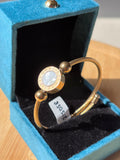 A gold-plated bangle bracelet with a round mother-of-pearl bezel and engraved brand lettering, displayed inside an open blue velvet jewelry box.