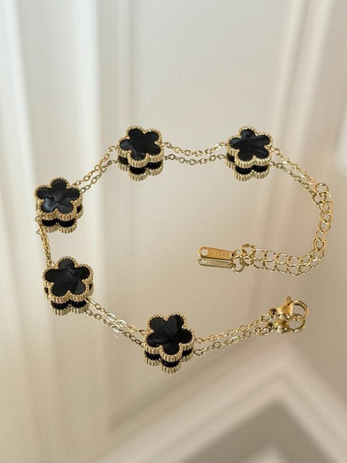 A gold chain bracelet featuring five alternating black flower-shaped charms with textured gold borders, displayed on a light cream background.