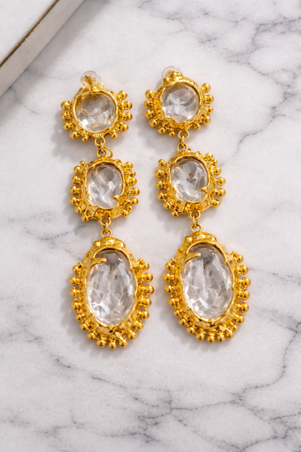 A pair of gold drop earrings with clear oval and round gemstones, displayed on a white marble surface.