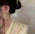 A close-up lifestyle shot of a model wearing a gold crescent-shaped hoop earring adorned with clear rhinestones. The model is wearing a pale yellow blouse and a pearl necklace, with her hair in a bun secured by a gold hair clip.