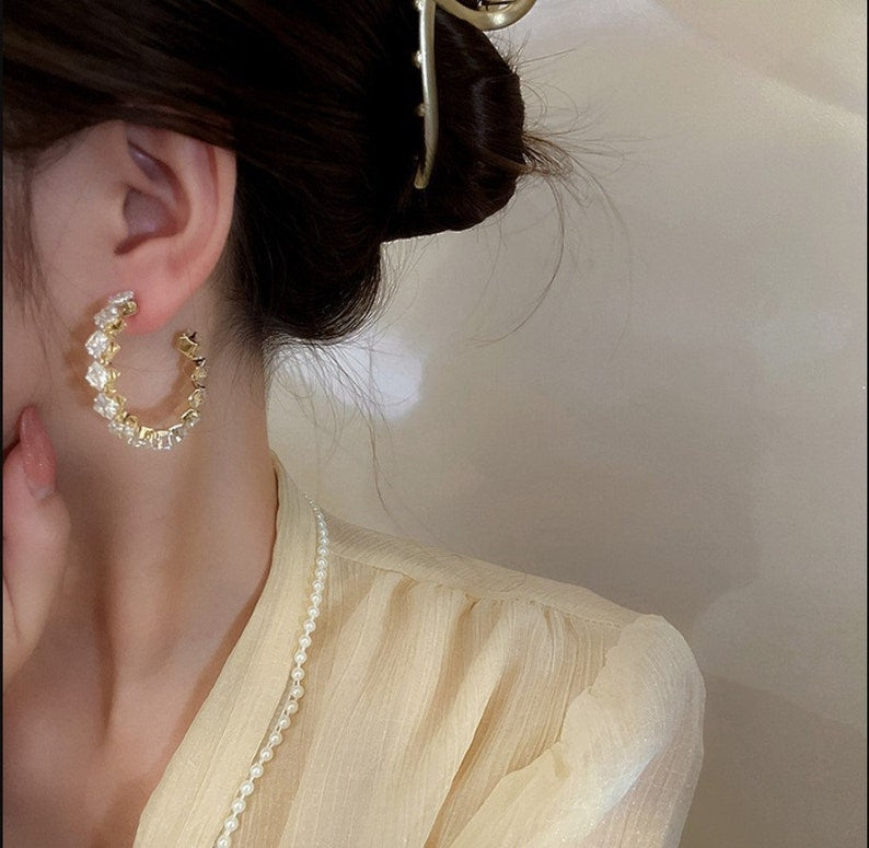 A close-up lifestyle shot of a model wearing a gold crescent-shaped hoop earring adorned with clear rhinestones. The model is wearing a pale yellow blouse and a pearl necklace, with her hair in a bun secured by a gold hair clip.