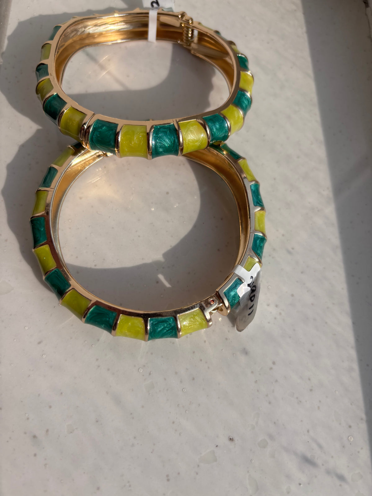 A pair of gold-plated bangle bracelets decorated with alternating green and yellow rectangular enamel tiles, resting on a light-colored surface.