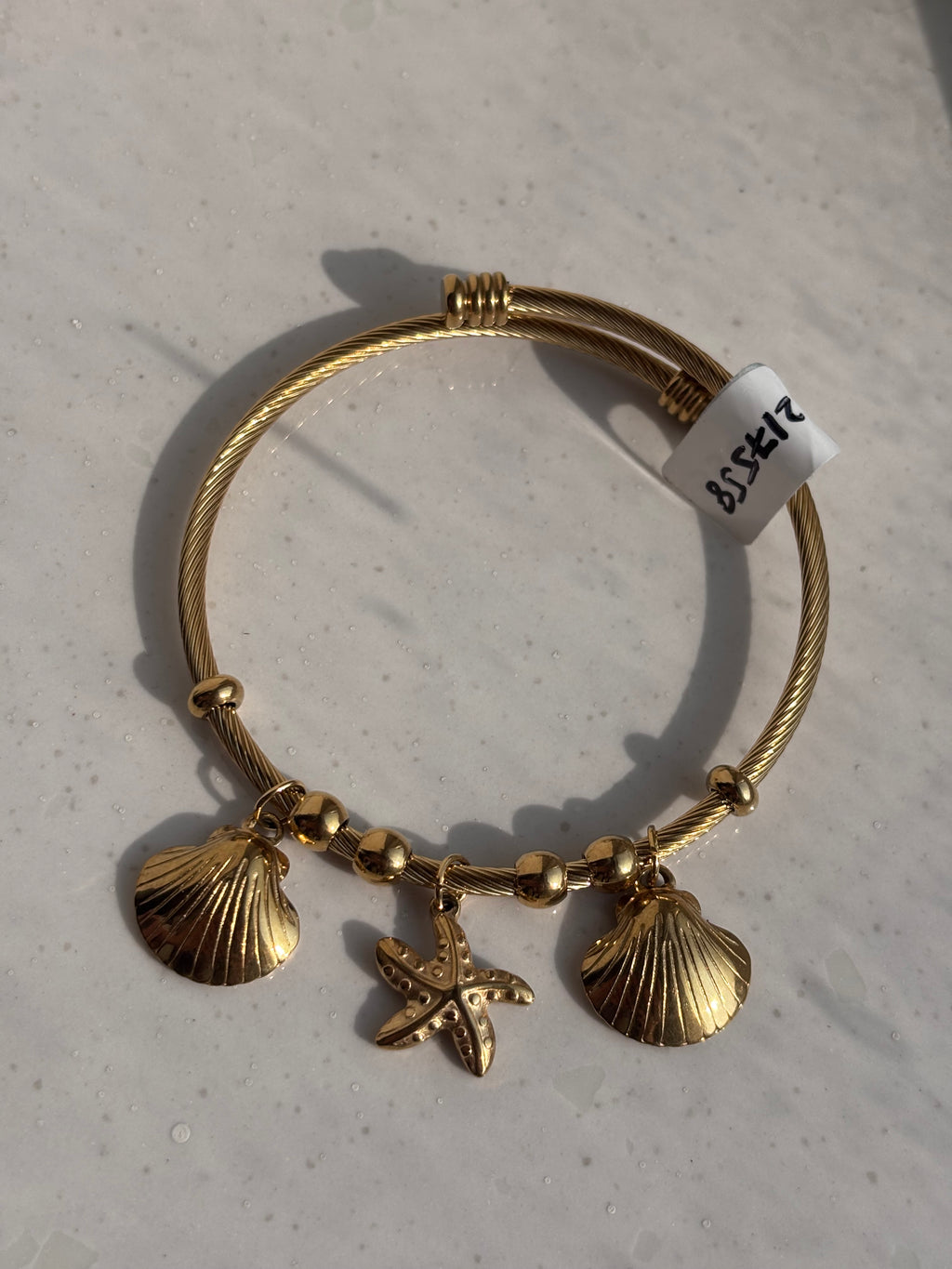 A gold-plated double-strand cable bangle bracelet with two shell charms and a starfish charm, resting on a light gray textured surface.