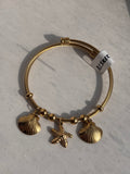 A gold-plated double-strand cable bangle bracelet with two shell charms and a starfish charm, resting on a light gray textured surface.