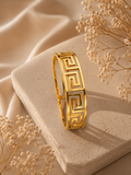 A gold bangle bracelet with a Greek key pattern, displayed on a textured beige stone block with dried baby's breath flowers and draped satin fabric.