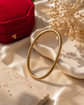 A polished gold oval bangle bracelet displayed on a cream ceramic dish, styled with a red velvet jewelry box and dried baby's breath flowers on a beige satin fabric background.