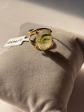 A gold ring with a large oval cabochon containing green and yellow marbled inclusions, displayed on a gray velvet cushion with a small white tag attached.