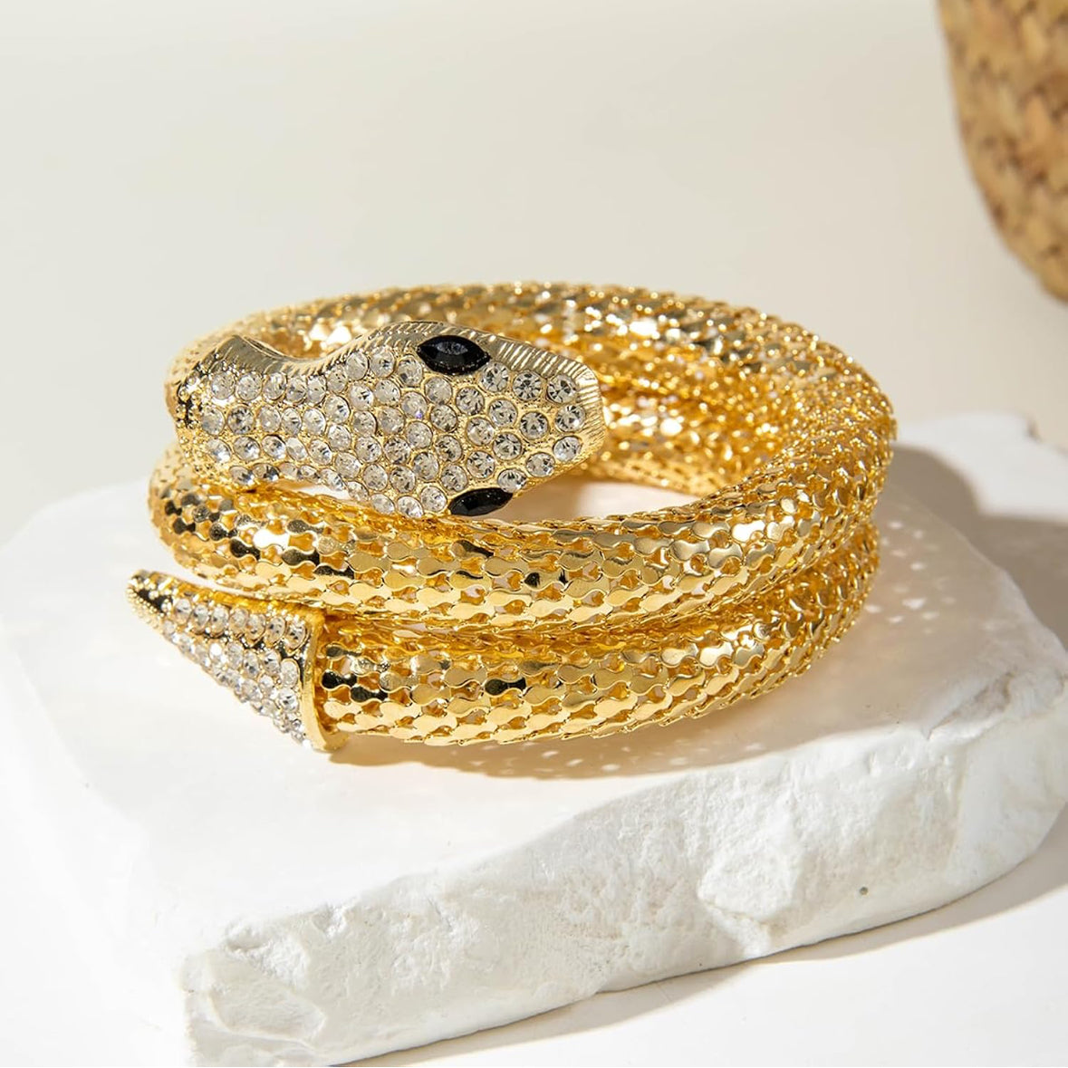 A gold snake cuff bracelet covered in clear rhinestones, featuring black rhinestone eyes, displayed on a white textured stone block.