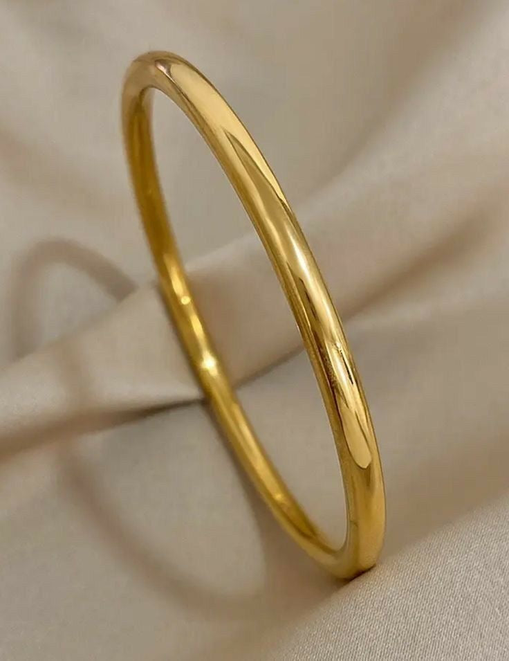 A polished yellow gold bangle bracelet with a smooth, rounded profile, resting on draped beige fabric.