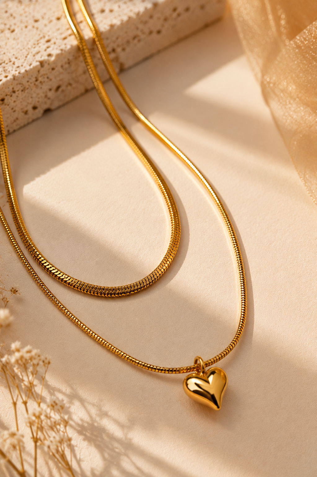A gold snake chain necklace with a polished heart-shaped pendant, styled on a warm beige surface with dried flowers and textured props.