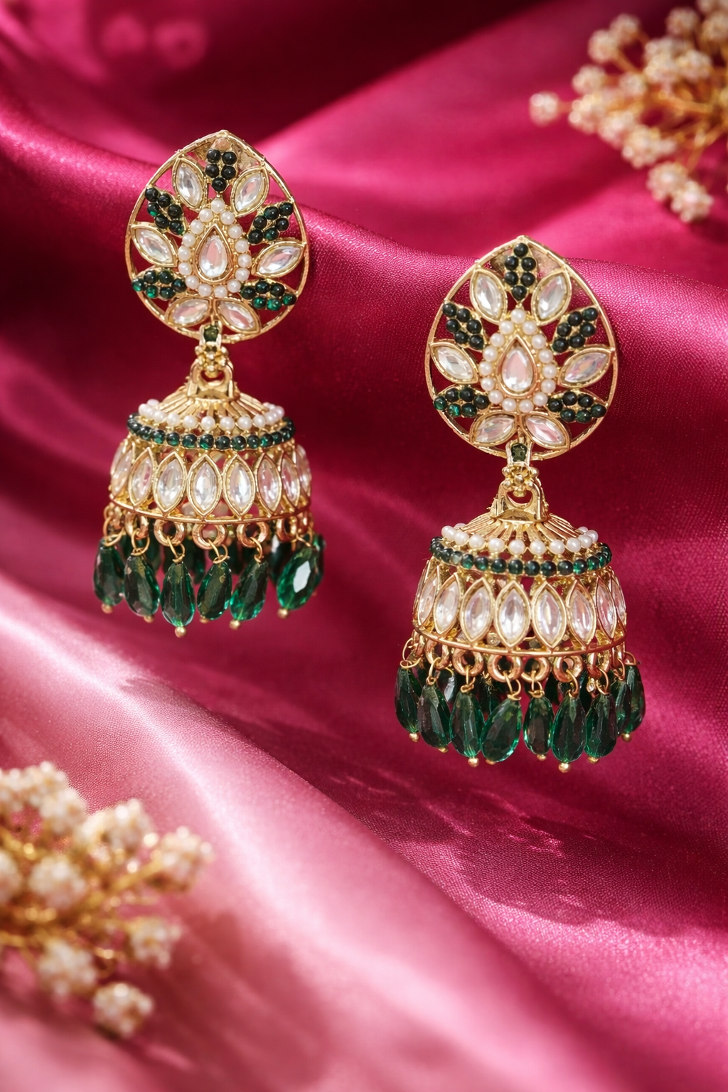 A pair of ornate gold-plated Indian jhumka earrings decorated with green stones, white stones, and dangling green teardrop crystals, displayed on a draped pink satin fabric.
