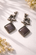 A pair of silver-toned drop earrings featuring a small dog figurine above a large square dark brown stone, displayed on a textured white surface with soft shadows and small white flowers.