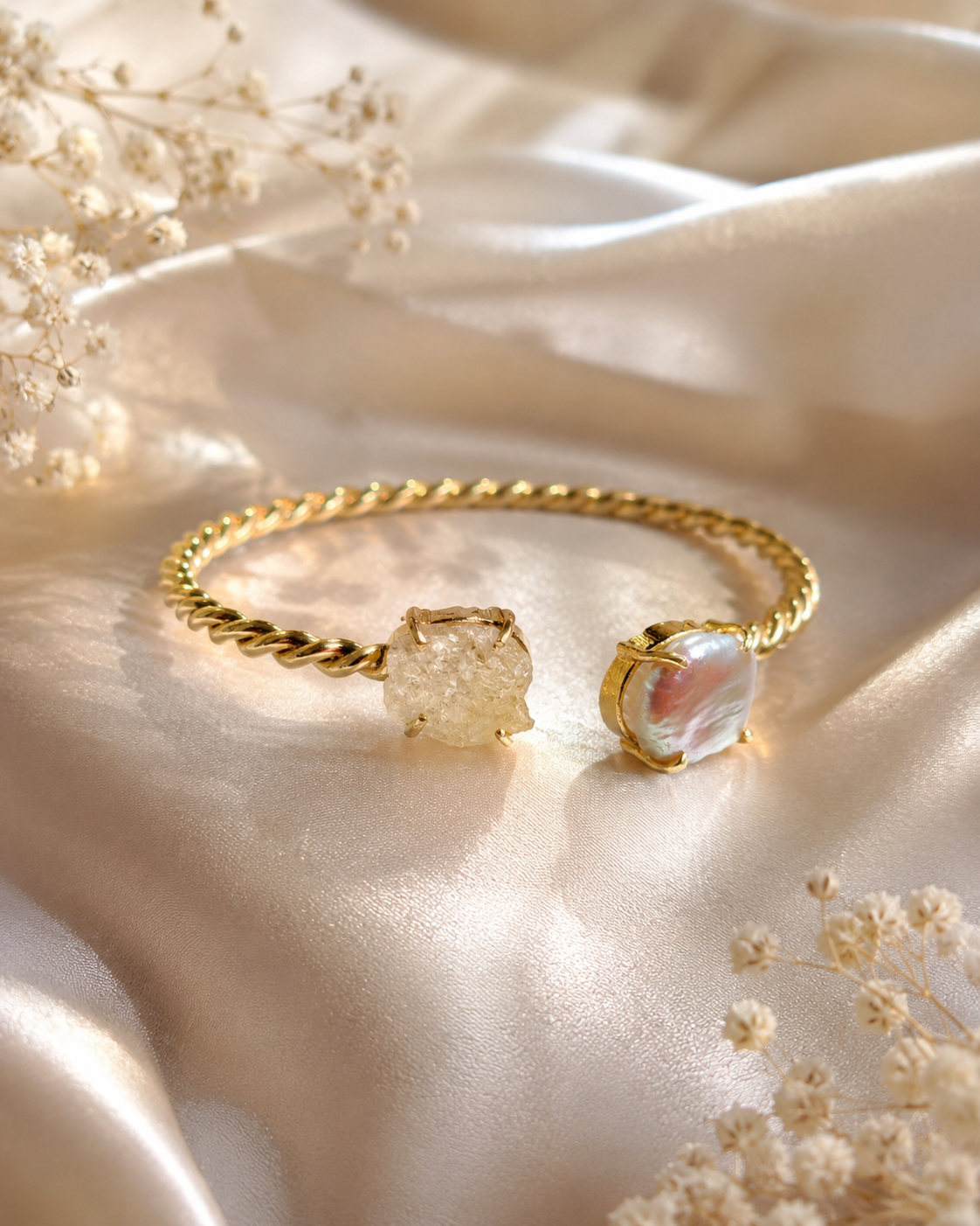 A gold twisted-band cuff bracelet with two textured stone charms, displayed on draped champagne satin fabric with dried baby's breath flowers.