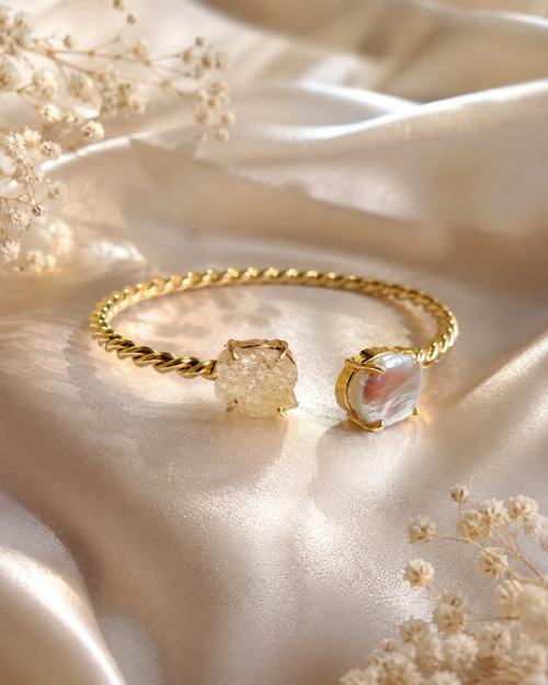 A gold twisted-band cuff bracelet with two textured stone charms, displayed on draped champagne satin fabric with dried baby's breath flowers.