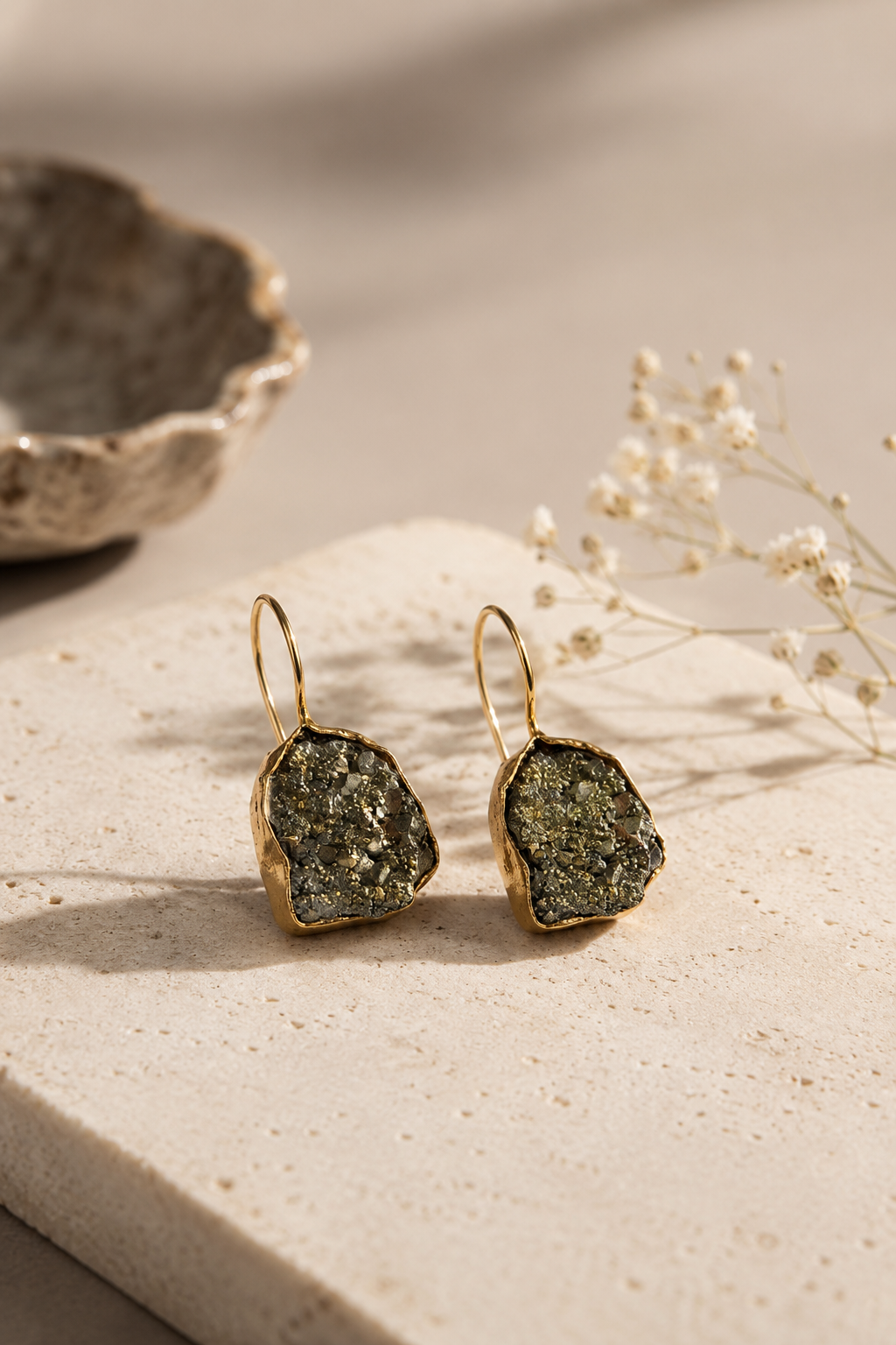 A pair of gold-tone drop earrings with irregular pyrite stone settings, displayed on a light beige stone slab with dried flowers and a ceramic bowl in the background.
