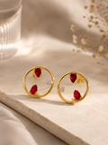 A pair of gold circular stud earrings with red teardrop gemstones and a small clear accent stone, displayed on a beige textured surface with soft warm lighting.