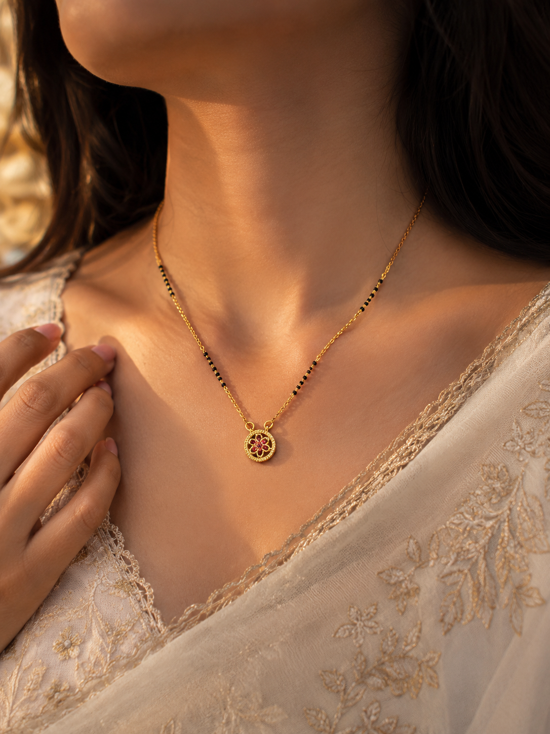 A close-up lifestyle shot of a woman wearing a delicate gold necklace with a circular pendant featuring a red floral design, styled with a cream-colored lace garment.