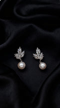 A pair of silver-toned drop earrings featuring a leaf-shaped cluster of clear stones and a single white pearl dangling below, displayed on a draped black satin fabric background.