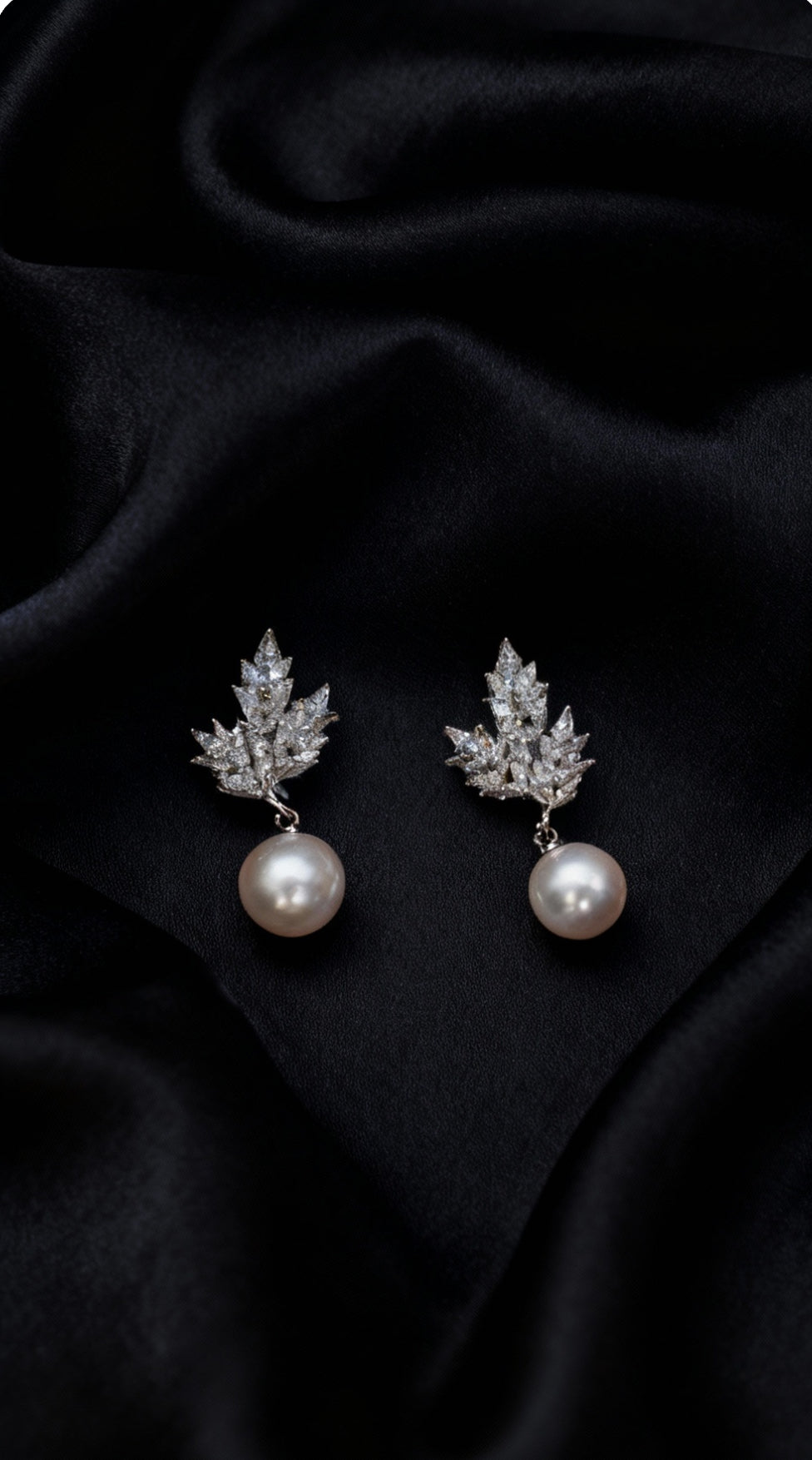 A pair of silver-toned drop earrings featuring a leaf-shaped cluster of clear stones and a single white pearl dangling below, displayed on a draped black satin fabric background.