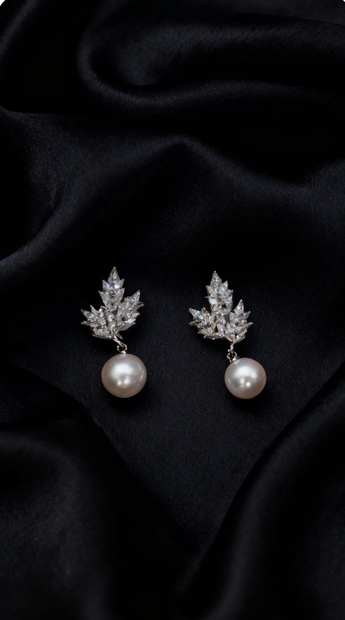 A pair of silver-toned drop earrings featuring a leaf-shaped cluster of clear stones and a single white pearl dangling below, displayed on a draped black satin fabric background.