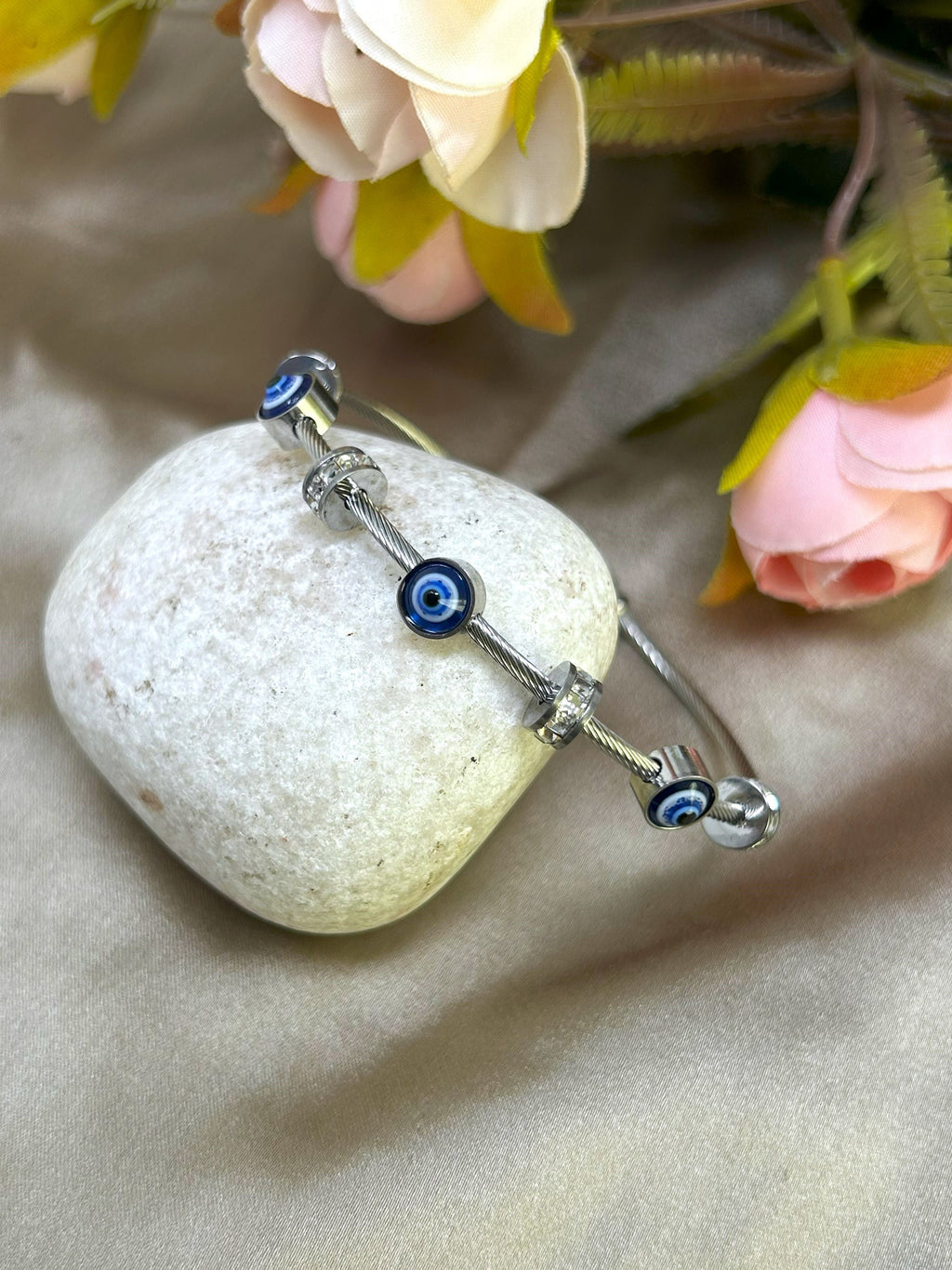 A silver-toned cuff bracelet with blue evil eye charms, displayed on a smooth white stone with soft pink and cream flowers in the background.