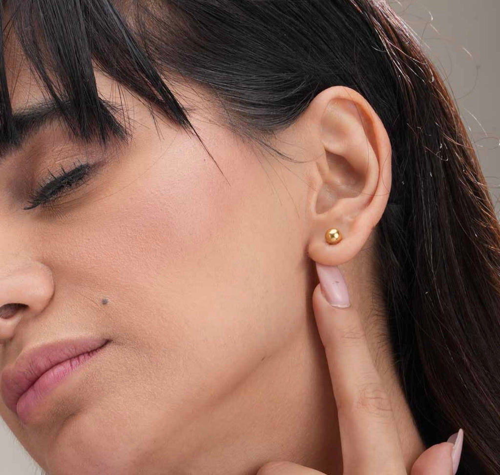A close-up of a woman's ear wearing a small gold stud earring, with her hand near her neck and her dark hair visible against a soft neutral background.