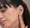 A close-up of a woman's ear wearing a small gold stud earring, with her hand near her neck and her dark hair visible against a soft neutral background.