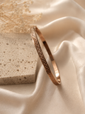 A rose gold bangle bracelet with an intricate geometric pattern, displayed on draped cream satin fabric next to a textured stone block.
