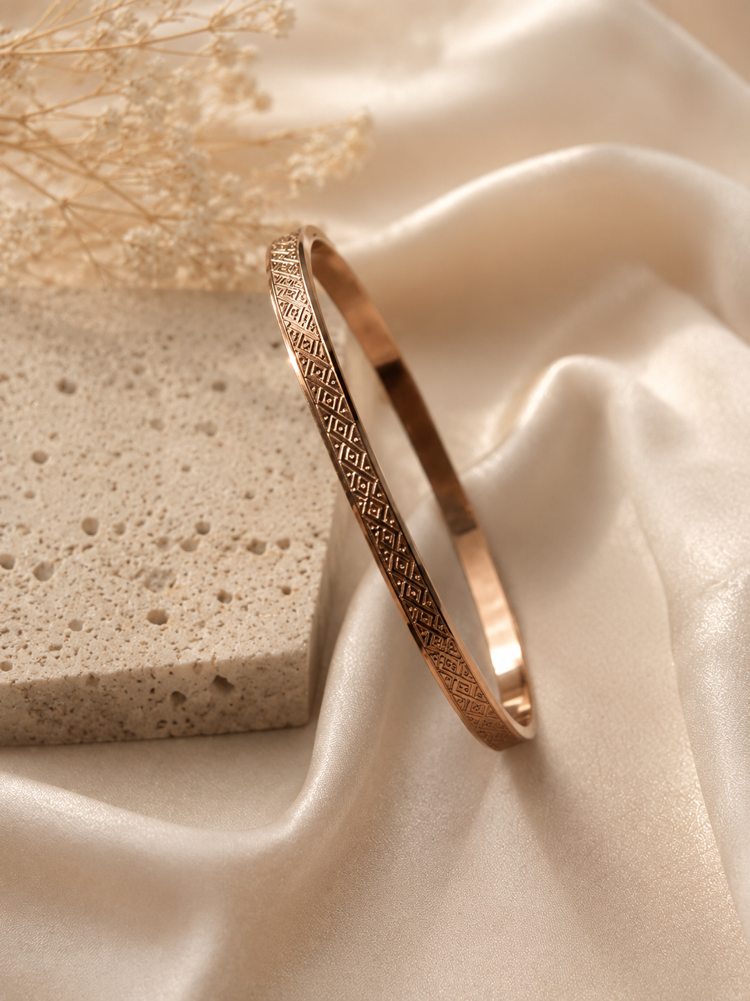 A rose gold bangle bracelet with an intricate geometric pattern, displayed on draped cream satin fabric next to a textured stone block.