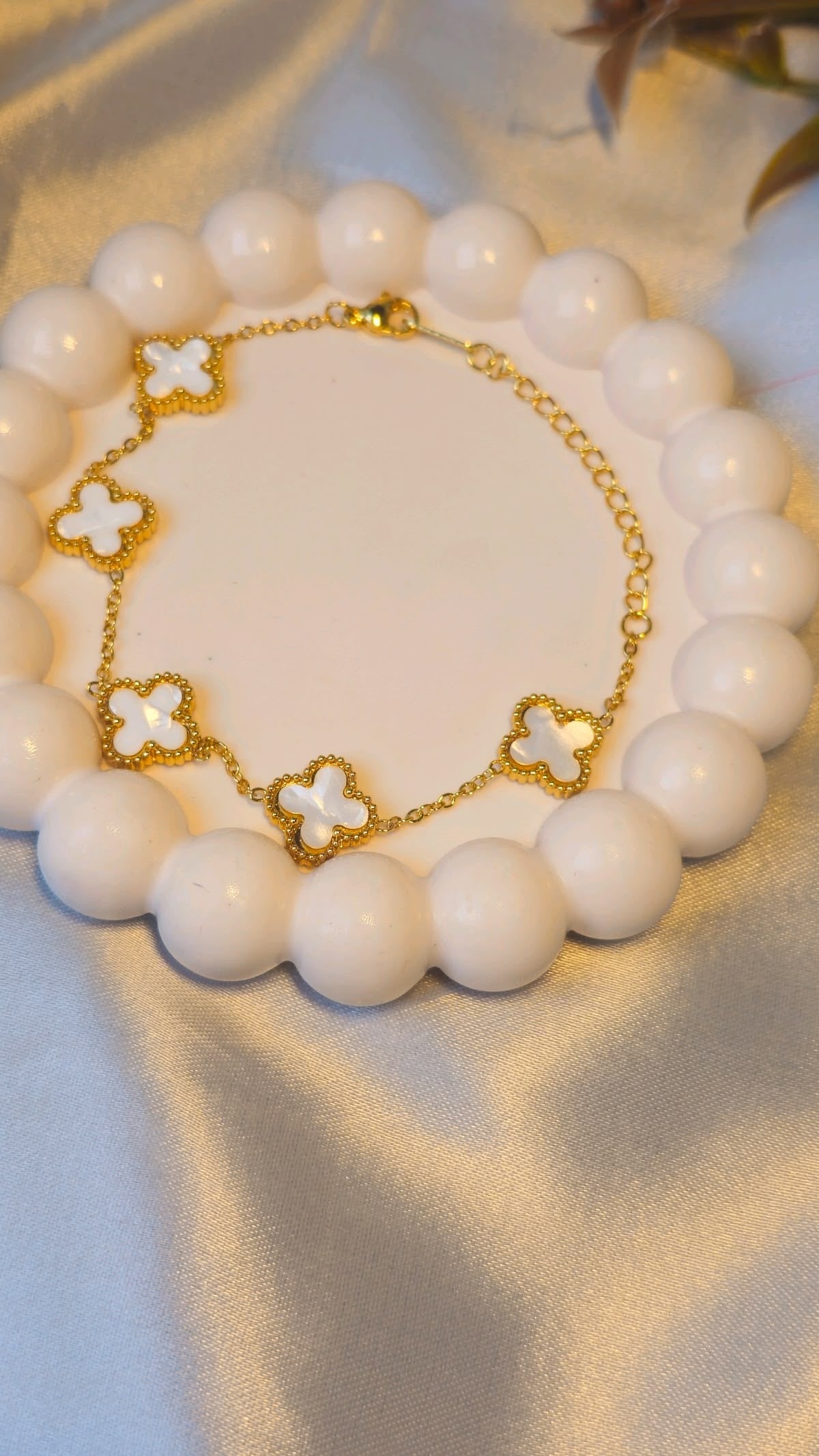 A gold chain bracelet with six white enamel four-leaf clover motifs, displayed on a circular white bead stand over a satin fabric background.