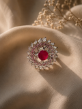 A circular silver-toned ring featuring a central round red gemstone surrounded by smaller pink and clear stones, displayed on draped beige fabric with dried floral elements in the background.