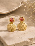 A pair of gold drop earrings with textured circular pendants and faceted brown gemstone studs, displayed on a beige stone surface with dried flowers in the background.