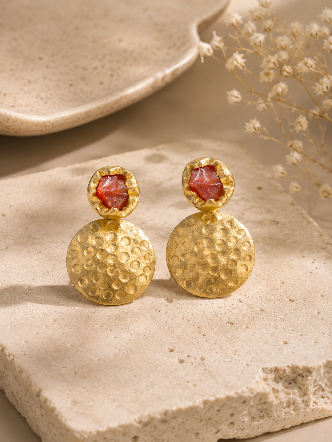 A pair of gold drop earrings with textured circular pendants and faceted brown gemstone studs, displayed on a beige stone surface with dried flowers in the background.