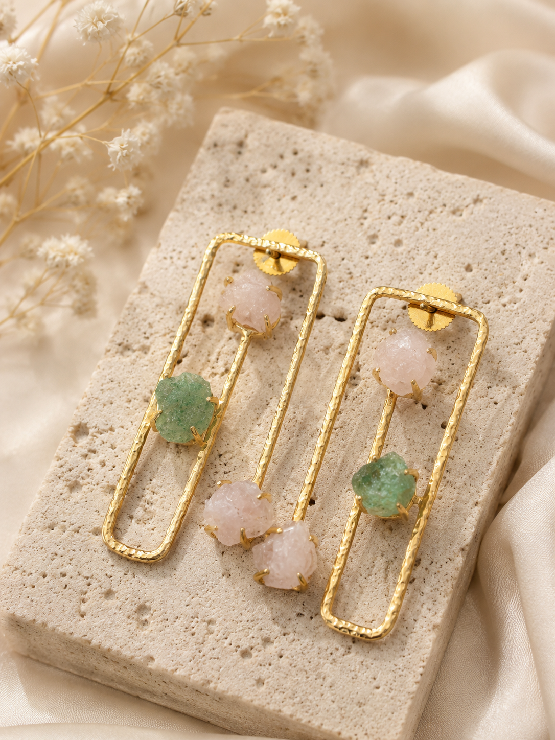 A pair of gold geometric drop earrings with pale pink and green gemstone accents, displayed on a textured stone block with soft cream fabric and dried flowers in the background.