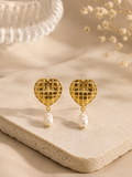 A pair of gold heart-shaped dangle earrings with a textured grid pattern and a single white pearl drop, displayed on a beige stone slab with dried flowers in the background.