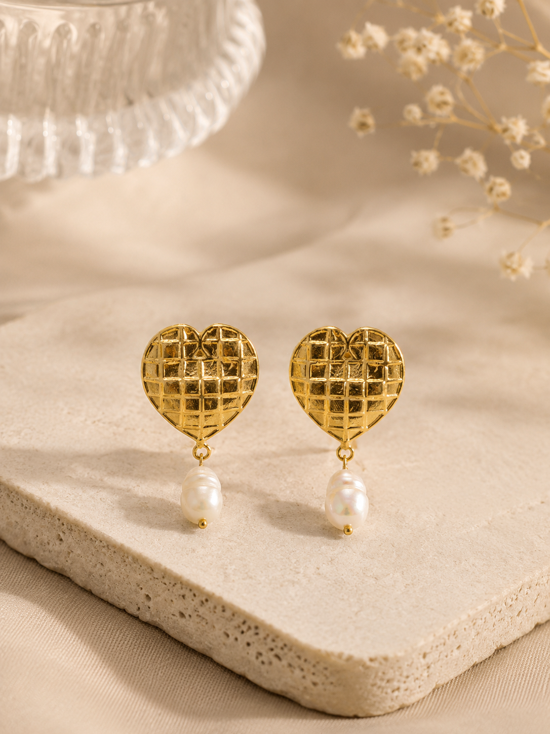 A pair of gold heart-shaped dangle earrings with a textured grid pattern and a single white pearl drop, displayed on a beige stone slab with dried flowers in the background.